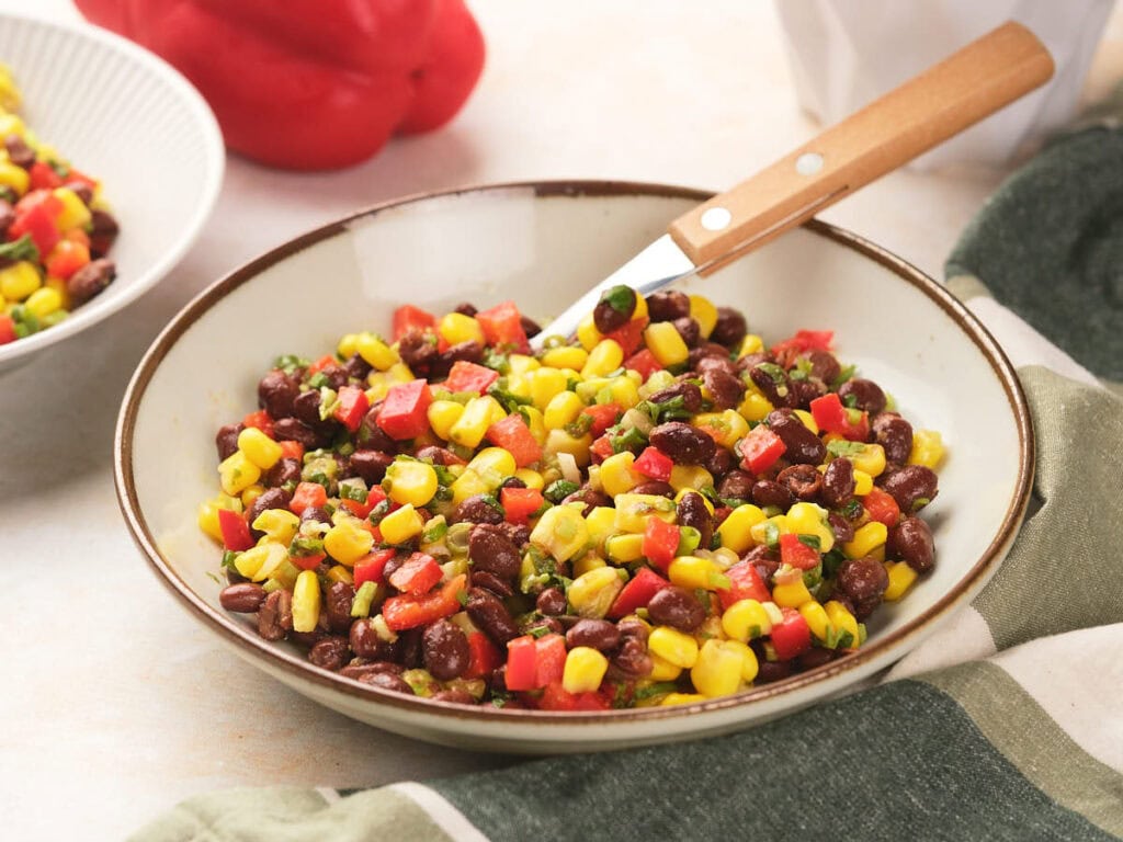 A bowl of black bean salad with diced red peppers and herbs, placed on a table with a fork resting in the bowl.