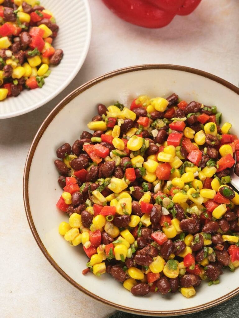 A bowl filled with a colorful black bean salad with a small portion in a separate dish nearby.