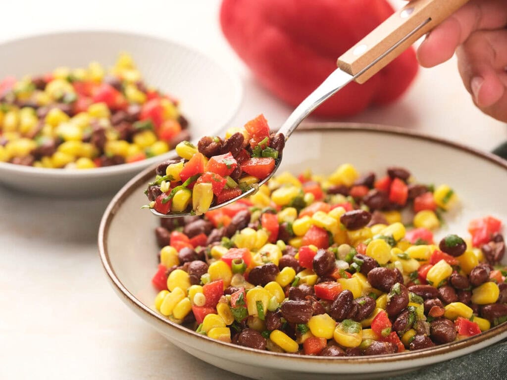 A spoon holds a mix of black bean salad filled with the same colorful bean salad.