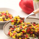 A spoonful of a colorful black bean salad with a bowl and red bell pepper in the background.