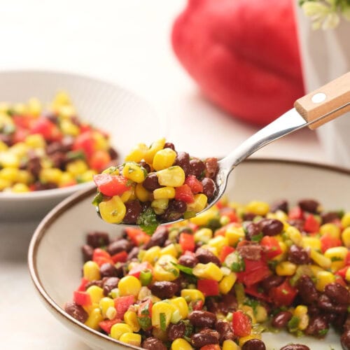 A spoonful of a colorful black bean salad with a bowl and red bell pepper in the background.