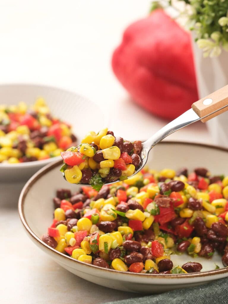 A spoonful of a colorful black bean salad with a bowl and red bell pepper in the background.