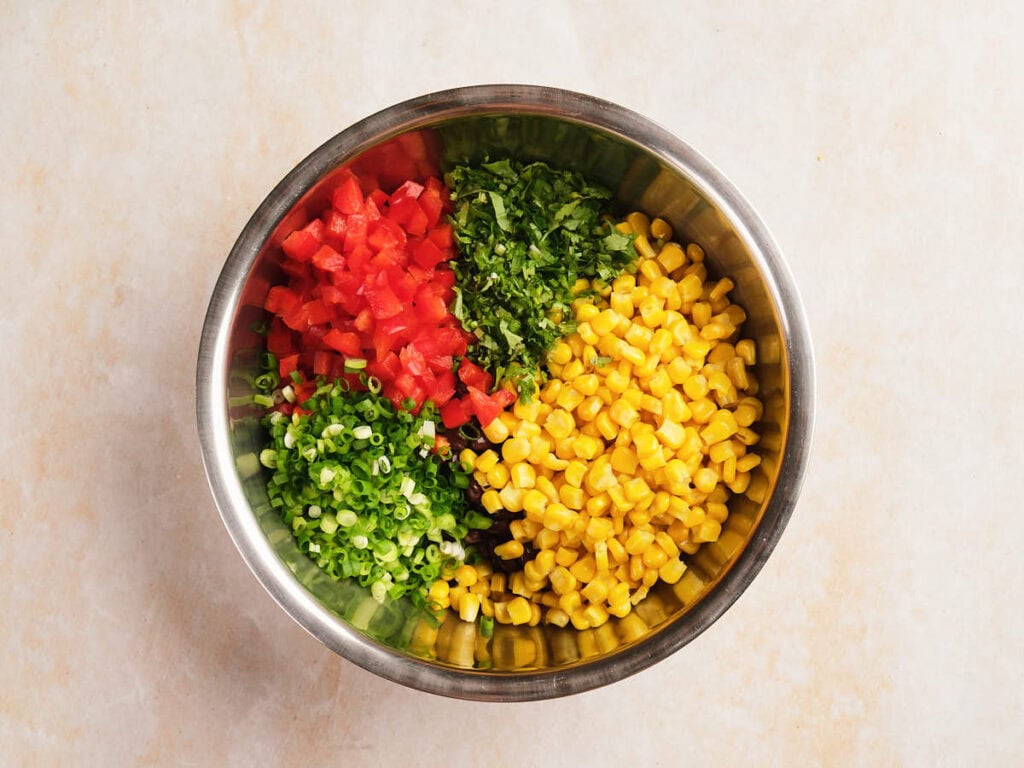 A metal bowl containing black bean salad ingredients are arranged in separate sections on a light surface.