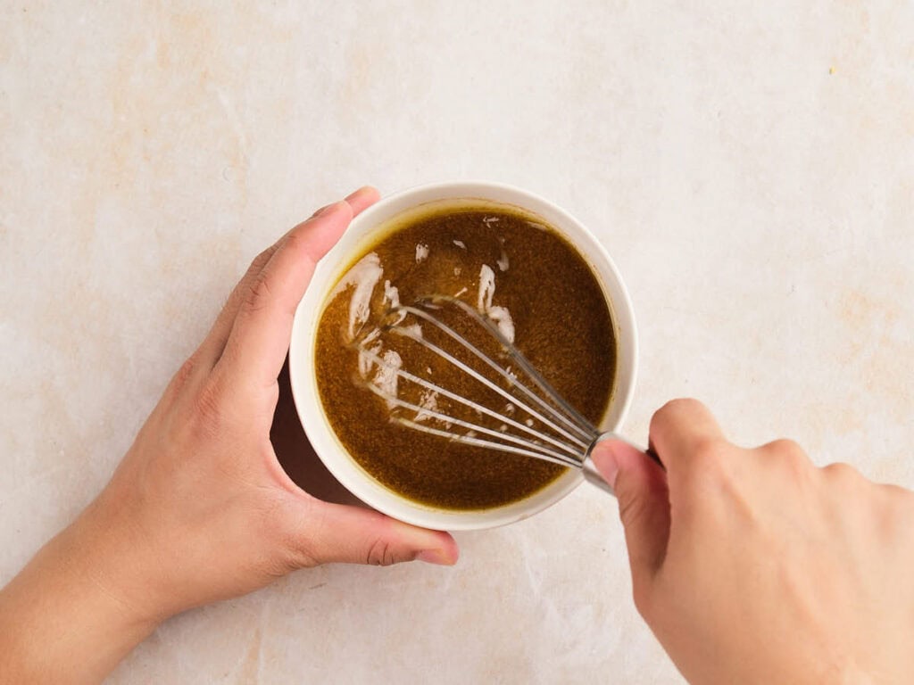 A person uses a whisk to mix a dark liquid in a white bowl, with one hand holding the bowl on a light-colored surface.