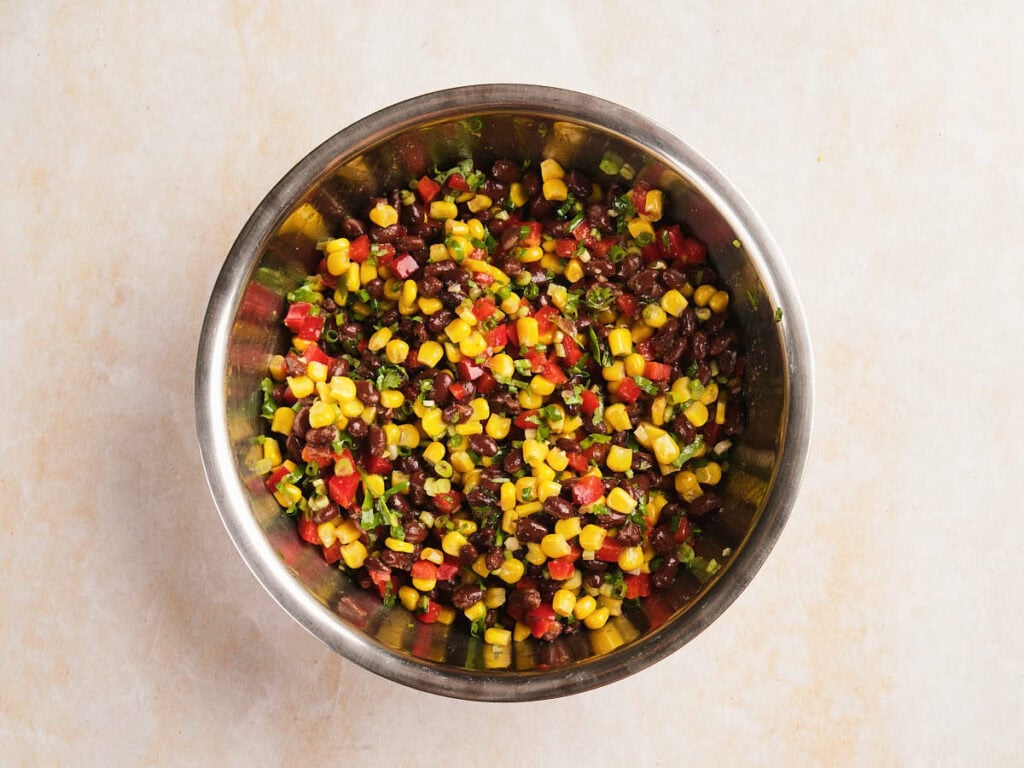 A stainless steel bowl filled with a colorful mixture of corn, diced tomatoes, chopped green herbs, and black beans on a light surface.