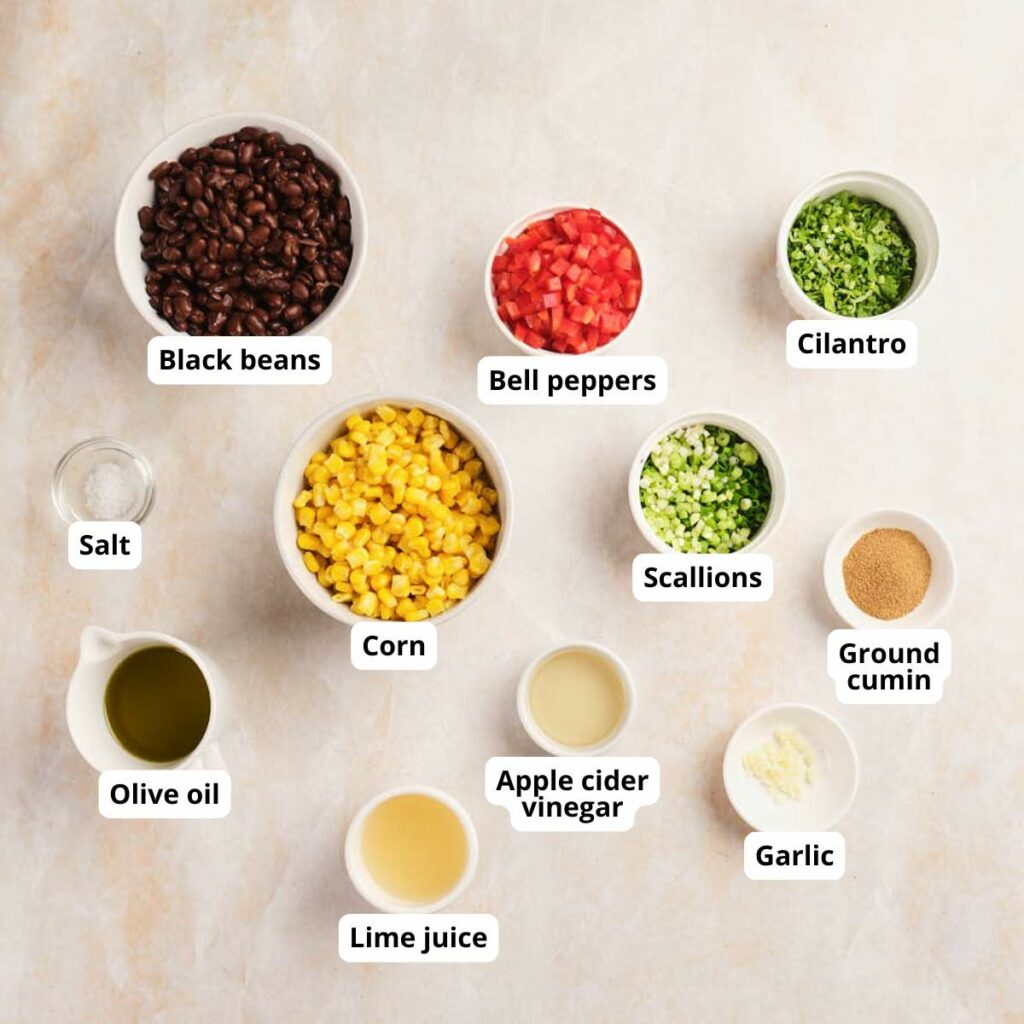 Various ingredients for a black bean salad recipe are arranged in bowls on a light surface.