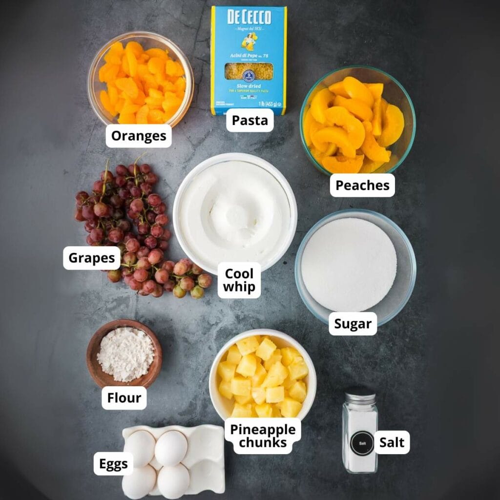 Top-view of labeled ingredients of fruit salad with cool whip recipe on a dark surface.