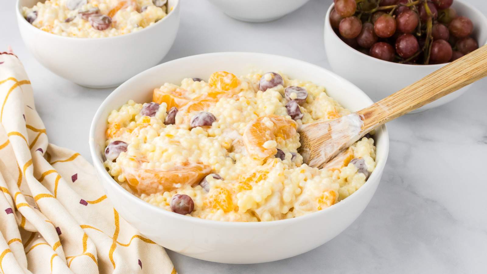A creamy fruit salad with cool whip next to a colorful table napkin and two small bowls on the background.