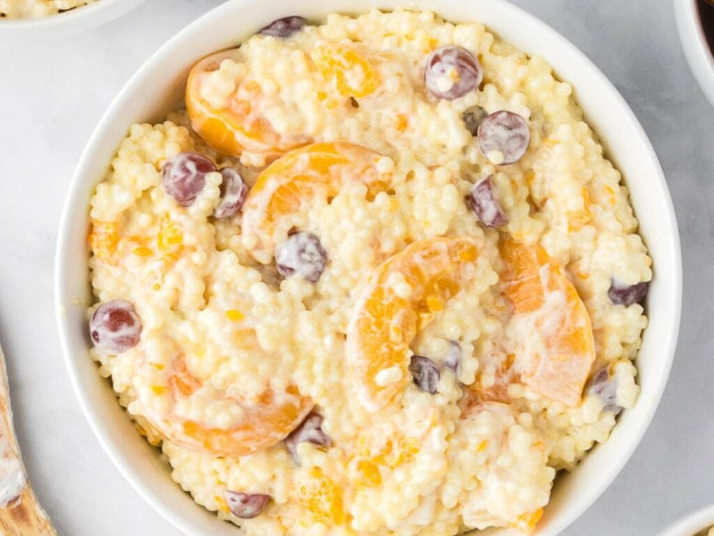 A close-up look of a creamy fruit salad with cool whip in a white bowl on a light surface.