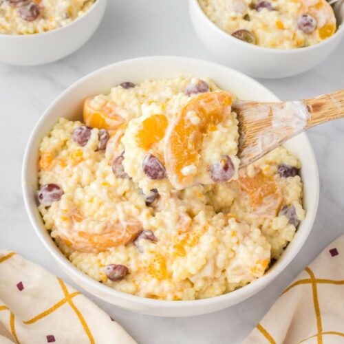 A white bowl of fruit salad with cool whip with a wooden spoon filled with the same salad.