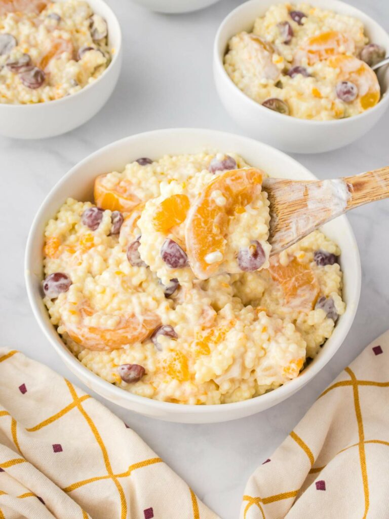 A white bowl of fruit salad with cool whip with a wooden spoon filled with the same salad.
