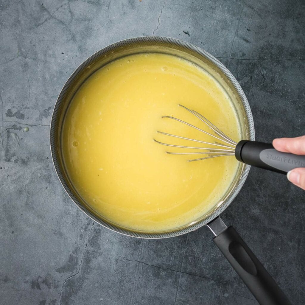A hand whisking a mixture reserved fruit juice with sugar, eggs, flour, and salt in a saucepan on a dark surface.