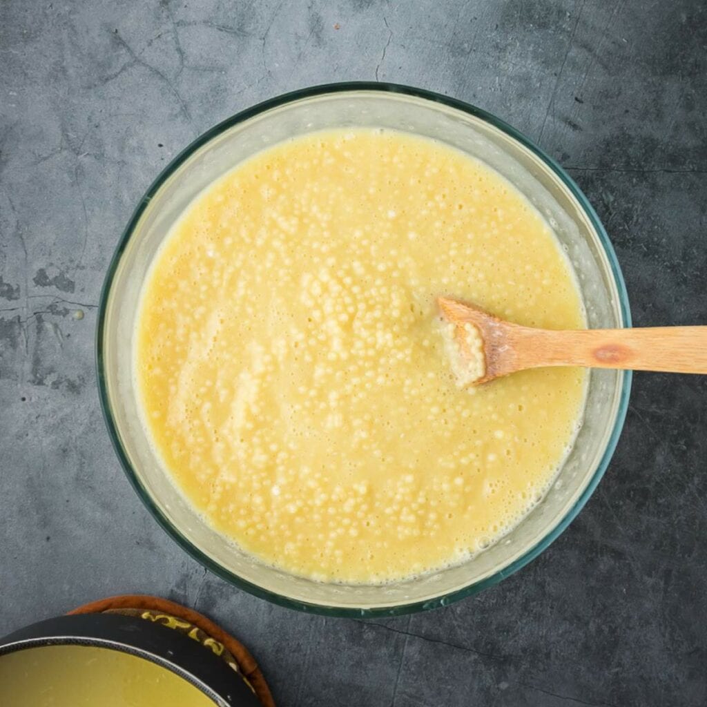 A glass bowl of cooked pasta with a wooden spoon inside next to a black small bowl with custard on a dark surface.