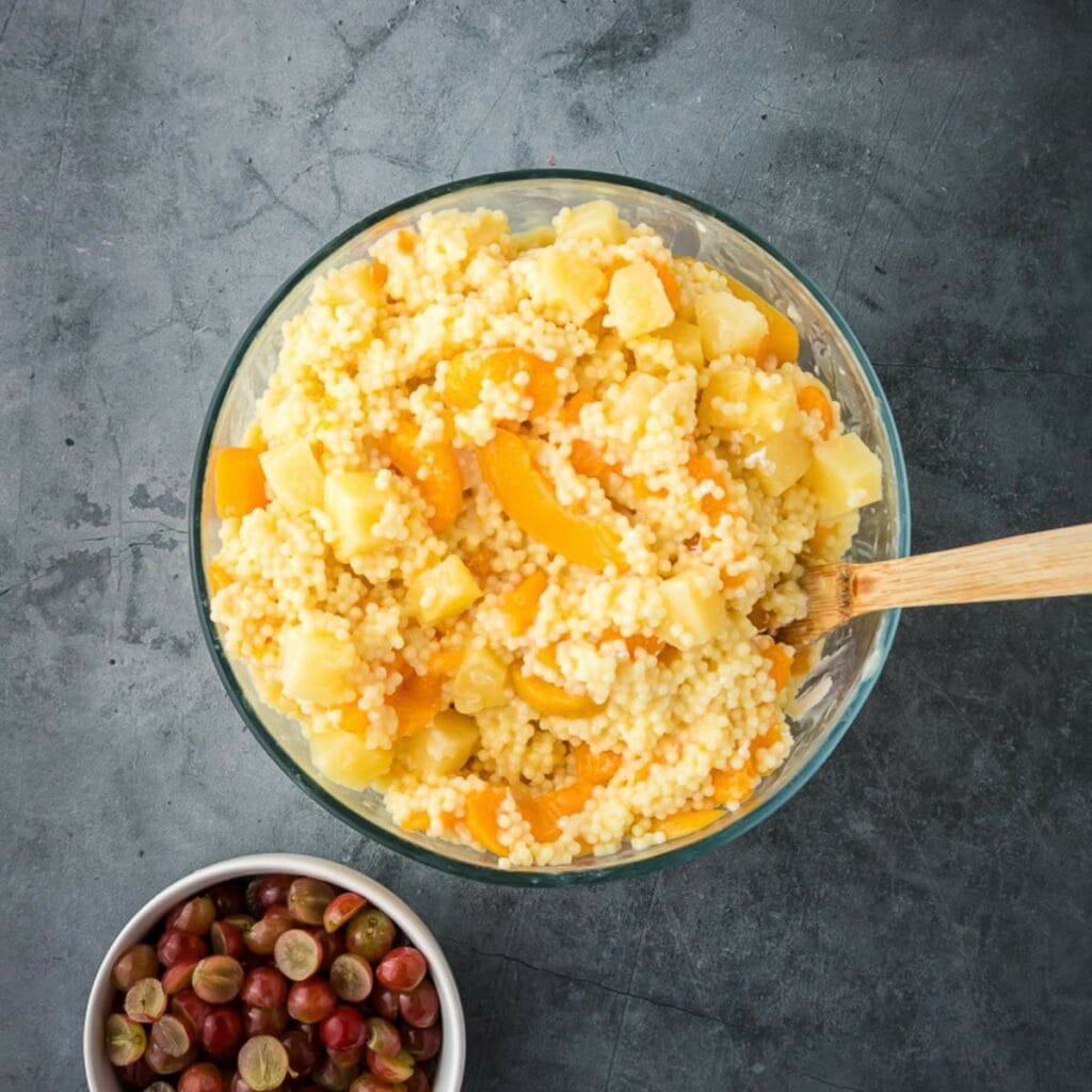 A mixture of cooked pasta and fruits in a glass bowl next to a bowl of grapes.