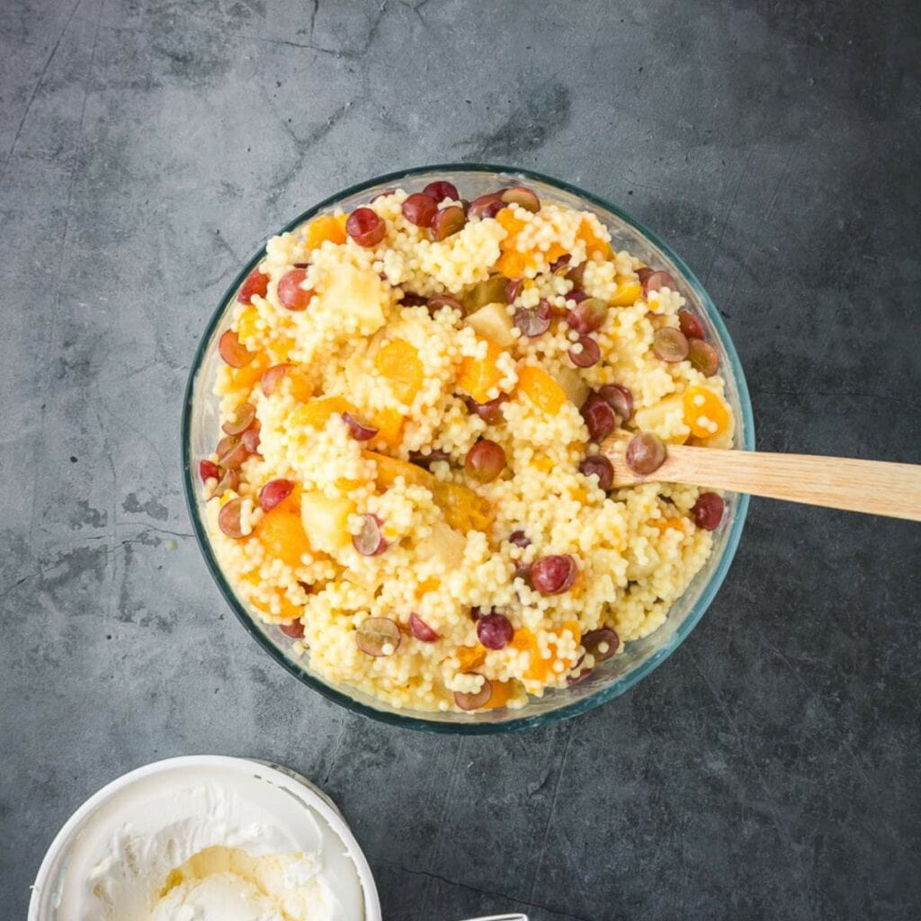 A big glass bowl with mix fruits and cooked pasta with a wooden spoon, next to a small bowl of a whip cream.