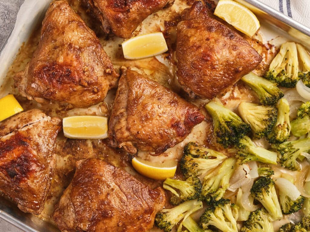 One pan chicken thighs with lemon wedges and roasted broccoli on a sheet pan.