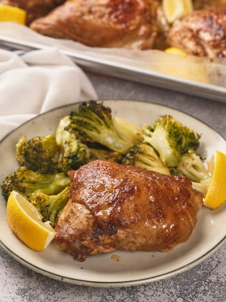 A plate with a cooked one pan chicken thighs, roasted broccoli, and two lemon wedges.