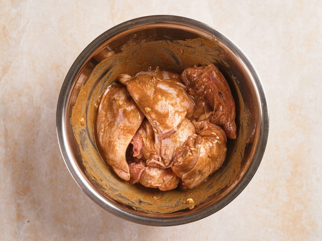 Raw chicken pieces coated in marinade sit in a stainless steel mixing bowl.