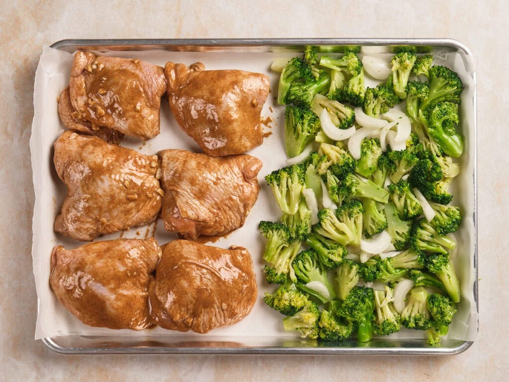 Sheet pan with four marinated chicken thighs on one side and chopped broccoli with onions on the other, ready to be cooked.