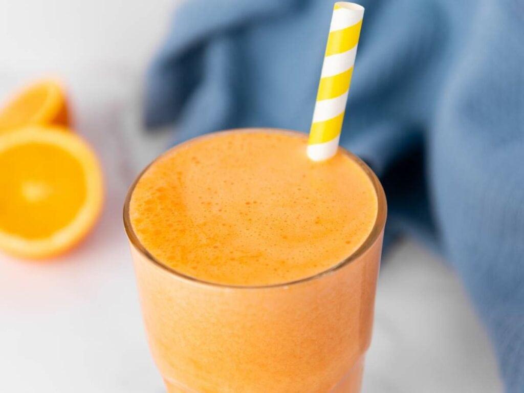 A glass of orange carrot juice with a yellow and white striped straw, with orange halves.