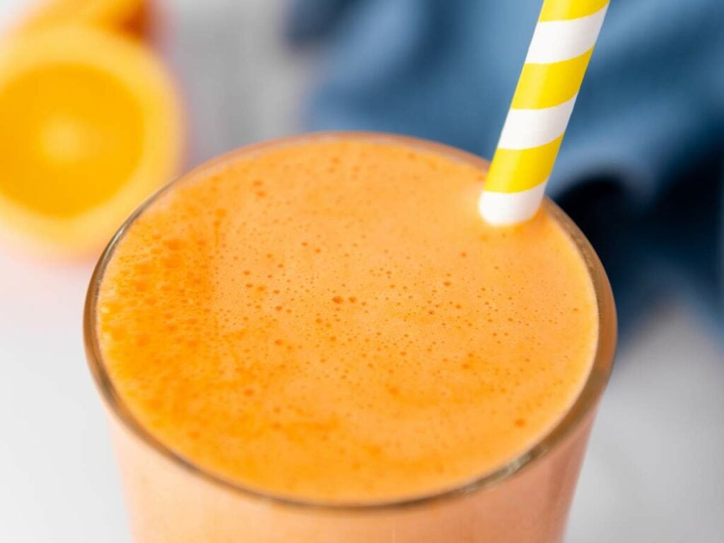 A close-up of an orange carrot juice in a glass with a yellow and white striped straw.
