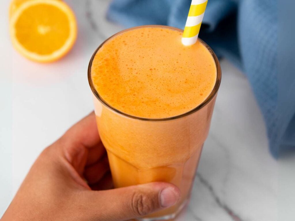A hand holds a glass of orange carrot juice with a yellow-and-white striped straw.