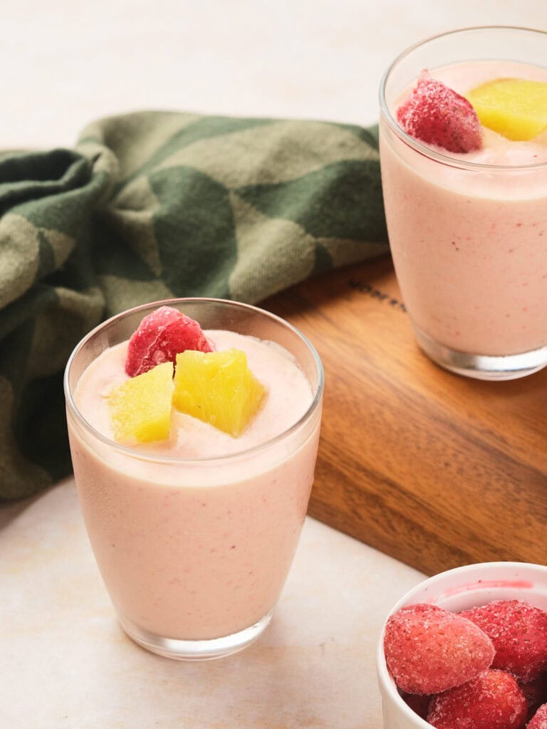 Two glasses of pineapple and strawberry smoothie topped with frozen strawberries and pineapple chunks are on a wooden board.