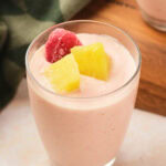 A glass of pineapple and strawberry smoothie topped with a frozen strawberry and pineapple chunks, placed on a light surface.