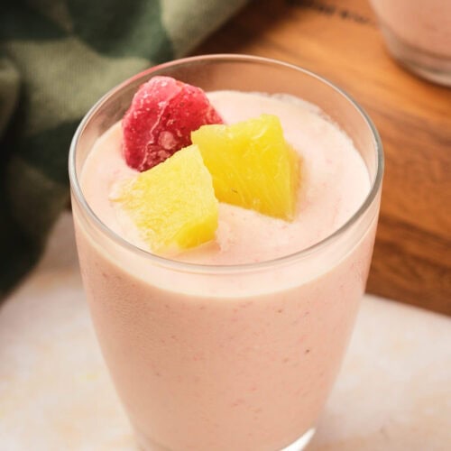 A glass of pineapple and strawberry smoothie topped with a frozen strawberry and pineapple chunks, placed on a light surface.
