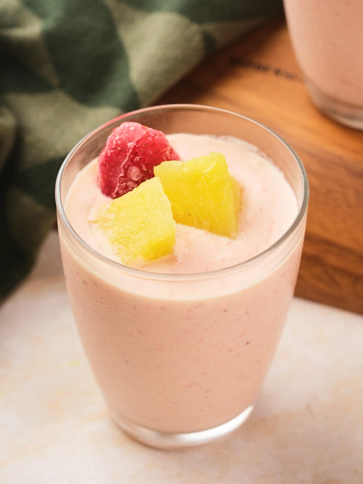 A glass of smoothie topped with a frozen strawberry and pineapple chunks, placed on a light surface.