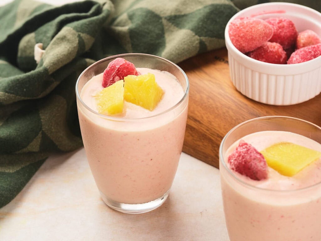 Two glasses of pineapple and strawberry smoothie topped with frozen strawberries and pineapple chunks, next to a bowl of frozen strawberries.