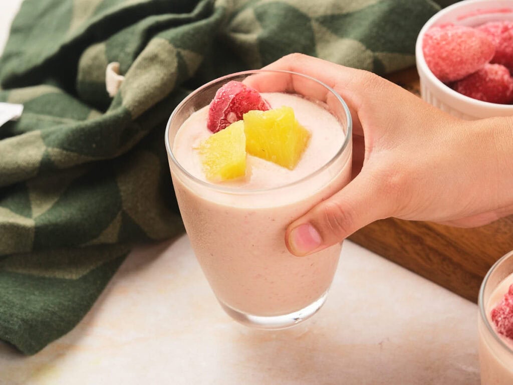 A hand holds a glass of pineapple and strawberry smoothie topped with frozen raspberries and pineapple chunks.