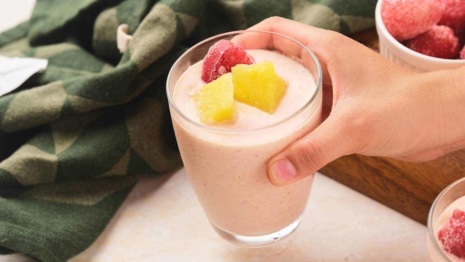 A hand holds a glass of pineapple and strawberry smoothie topped with pineapple chunks and strawberries.