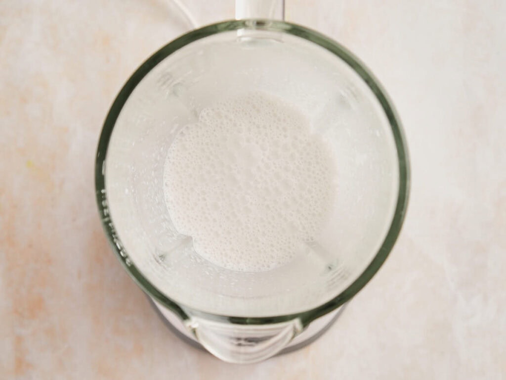 A top-down view of a blender containing coconut milk and white chocolate on a light-colored countertop.