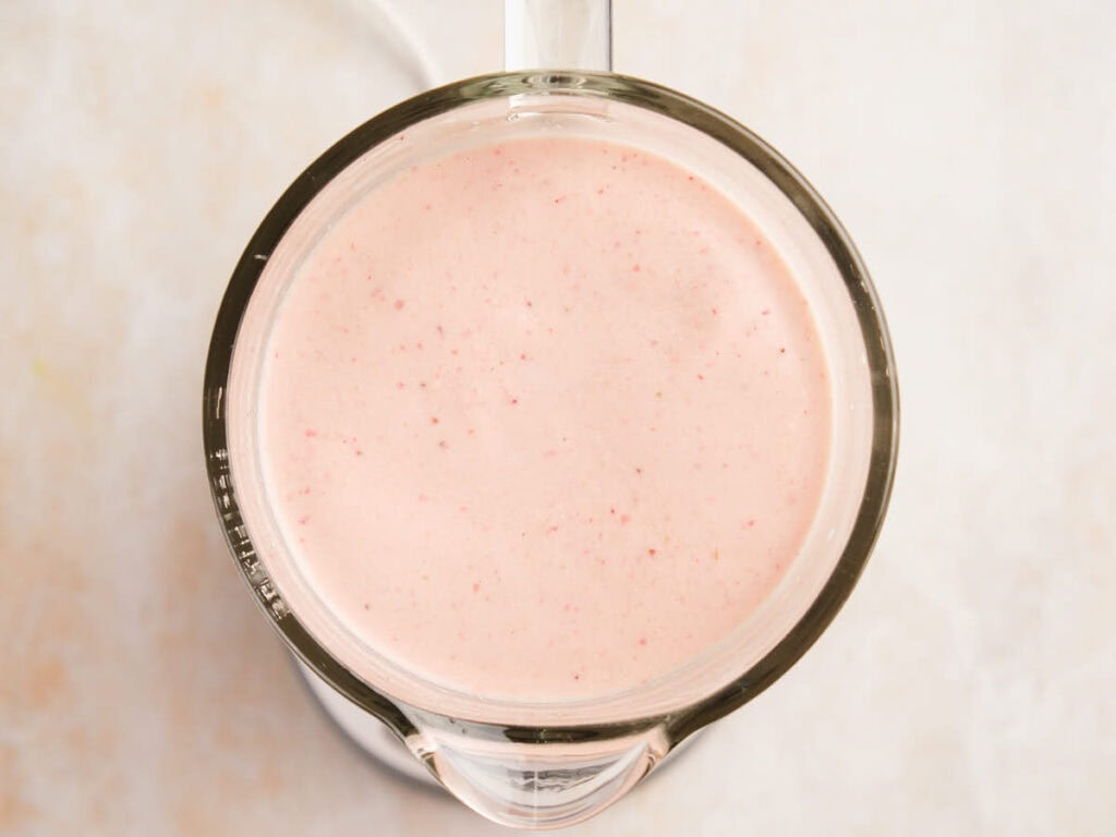 Overhead view of a blender filled with a creamy, light pink smoothie on a light-colored surface.