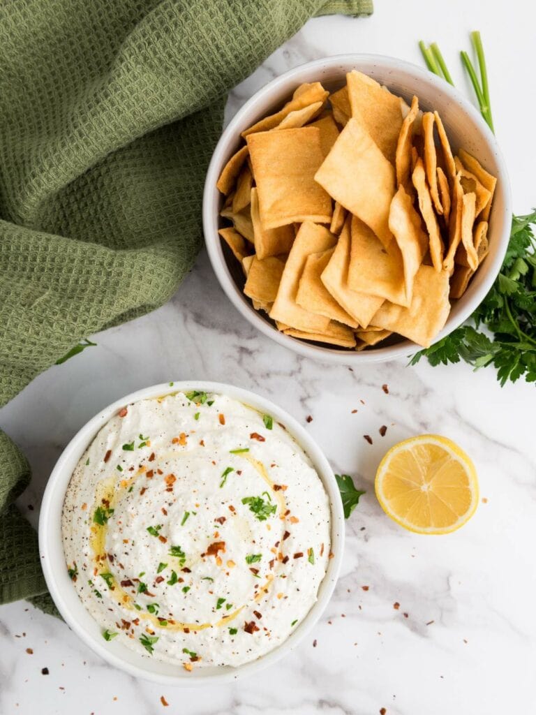 A bowl of chips next to a bowl of whipped feta cheese dip garnished with herbs and chili flakes, with a lemon half and green towel.