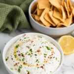 A bowl of whipped feta cheese dip garnished with herbs and chili flakes sits next to a bowl of chips.