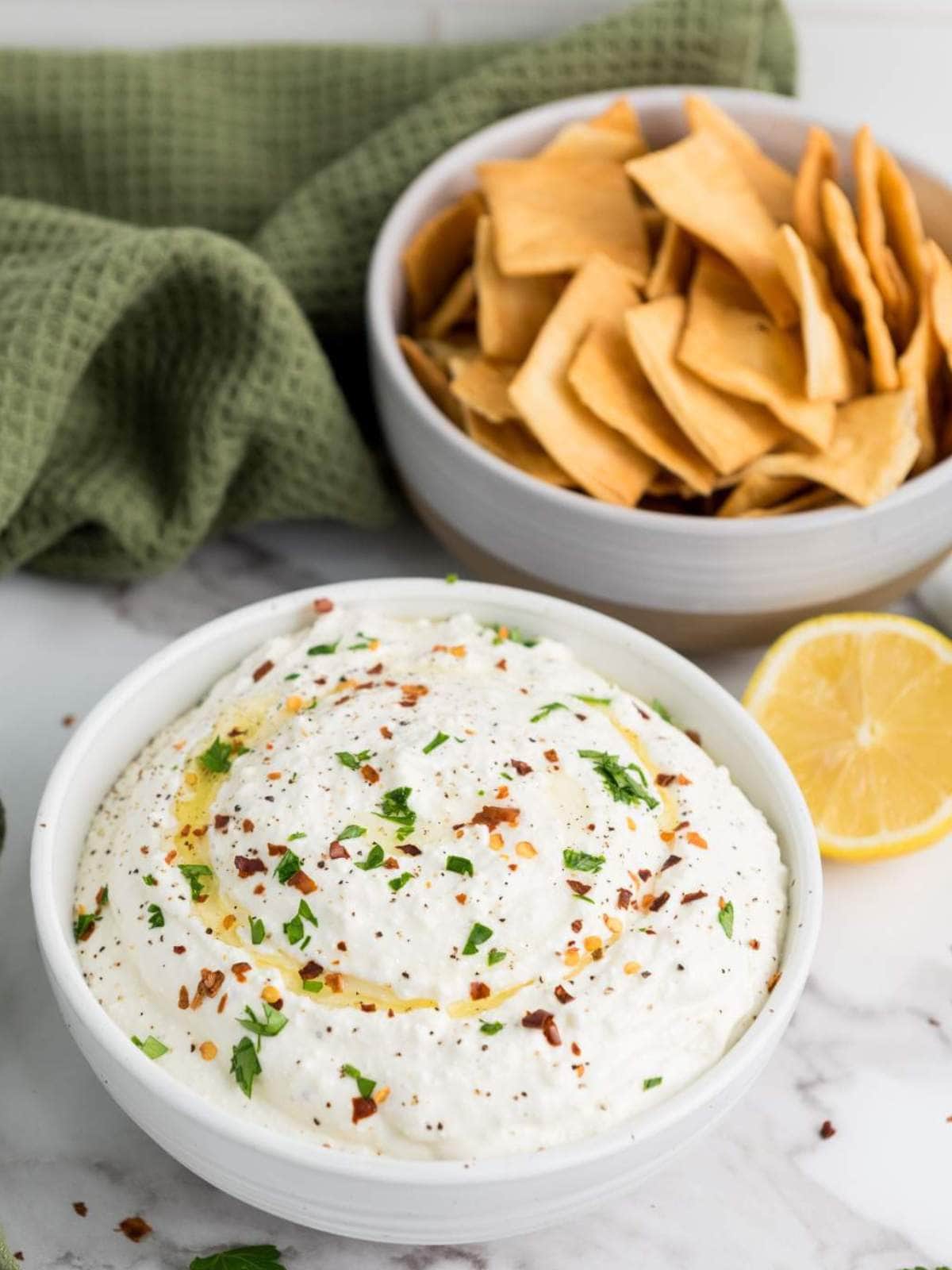 A bowl of whipped feta cheese dip garnished with herbs and chili flakes sits next to a bowl of chips.