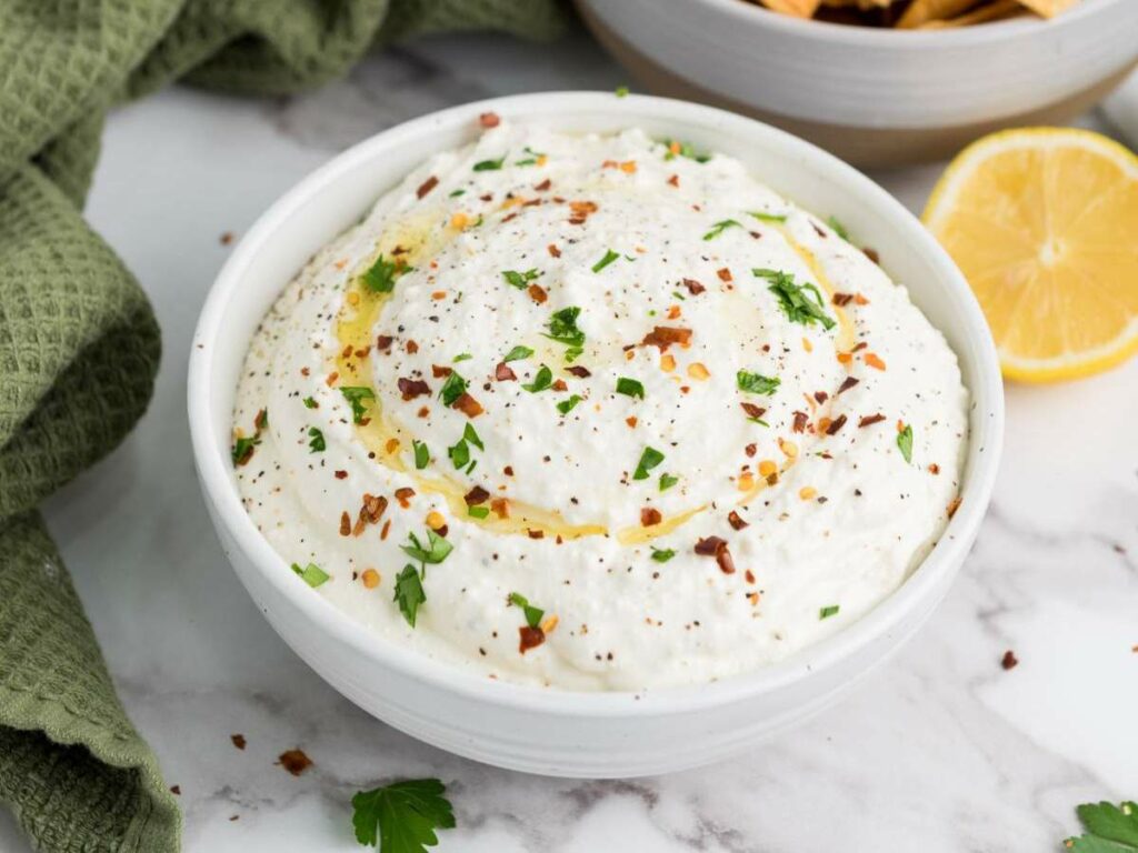 A white bowl filled with whipped feta cheese dip, garnished with olive oil, chopped herbs, and red pepper flakes.