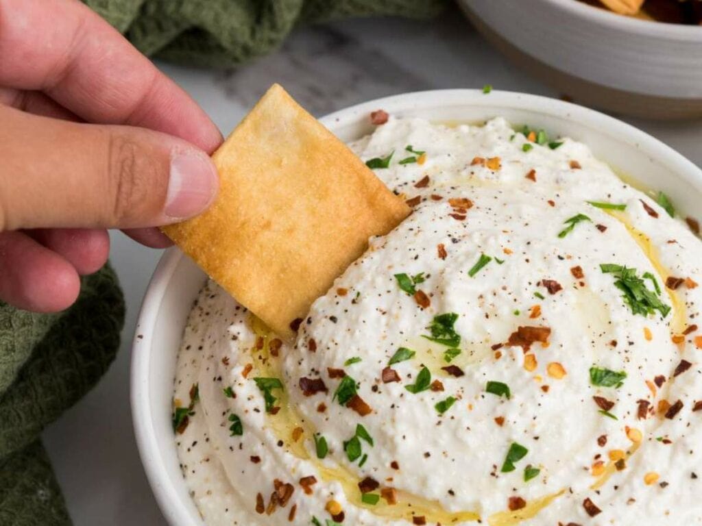 A hand dips a square cracker into a bowl of whipped feta cheese dip topped with herbs, pepper, and a drizzle of olive oil.