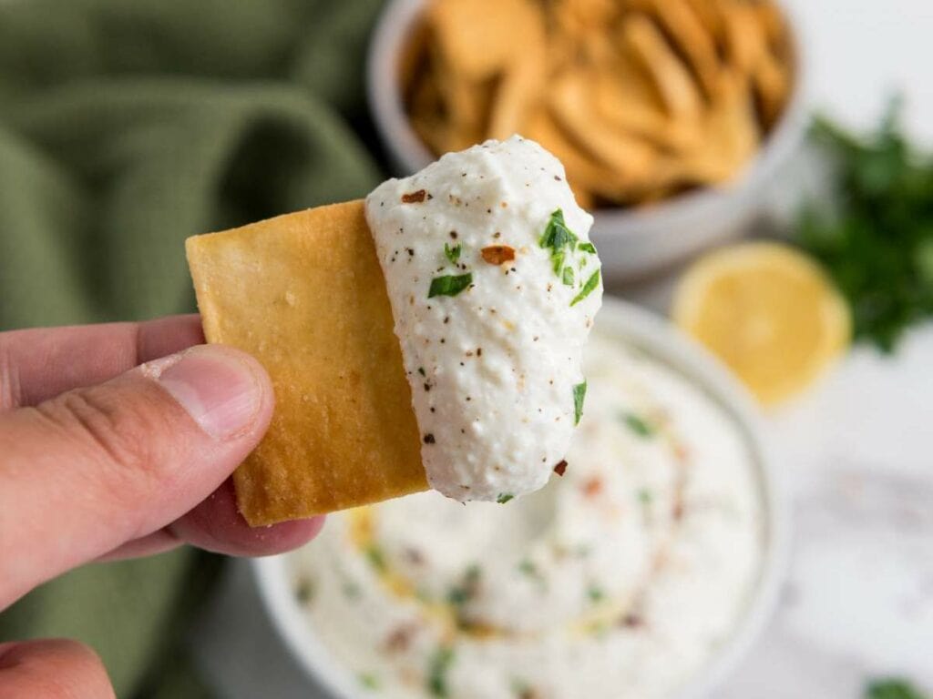 A hand holds a square cracker topped with a creamy feta cheese dip speckled with herbs and pepper.