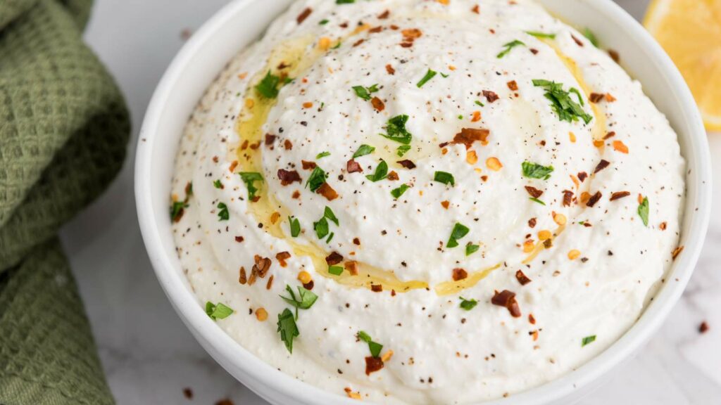 A bowl of creamy feta cheese dip garnished with chopped herbs, red pepper flakes, black pepper, and a drizzle of olive oil.