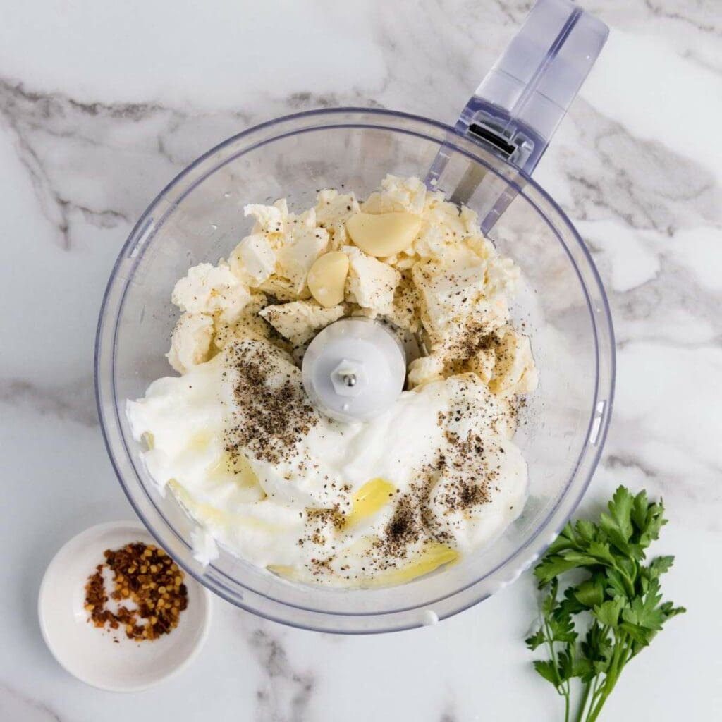 Food processor bowl containing yogurt, garlic, cheese, and black pepper on a marble surface.