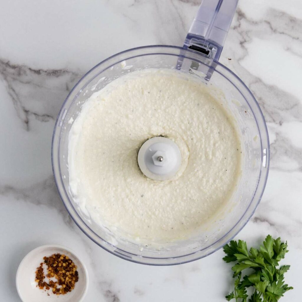 A food processor with blended white sauce on a marble surface, next to a small bowl of red pepper flakes and a sprig of parsley.