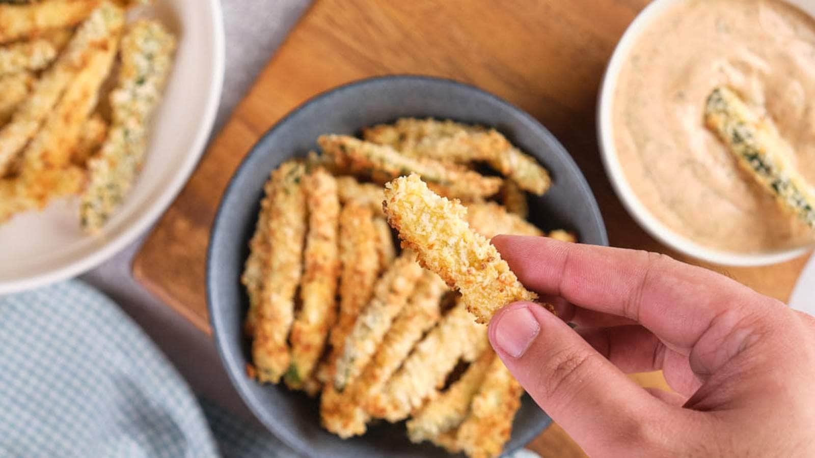 A hand holds a breaded zucchini fries with a bowl of dipping sauce nearby.