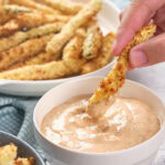 A hand dips a breaded zucchini fry into a bowl of creamy dipping sauce, with more zucchini fries on a plate in the background.