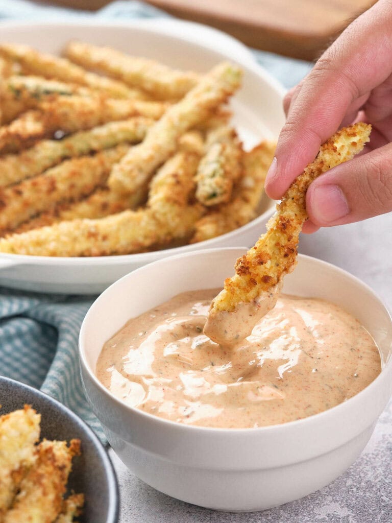 A hand dips a breaded zucchini fry into a bowl of creamy dipping sauce, with more zucchini fries on a plate in the background.