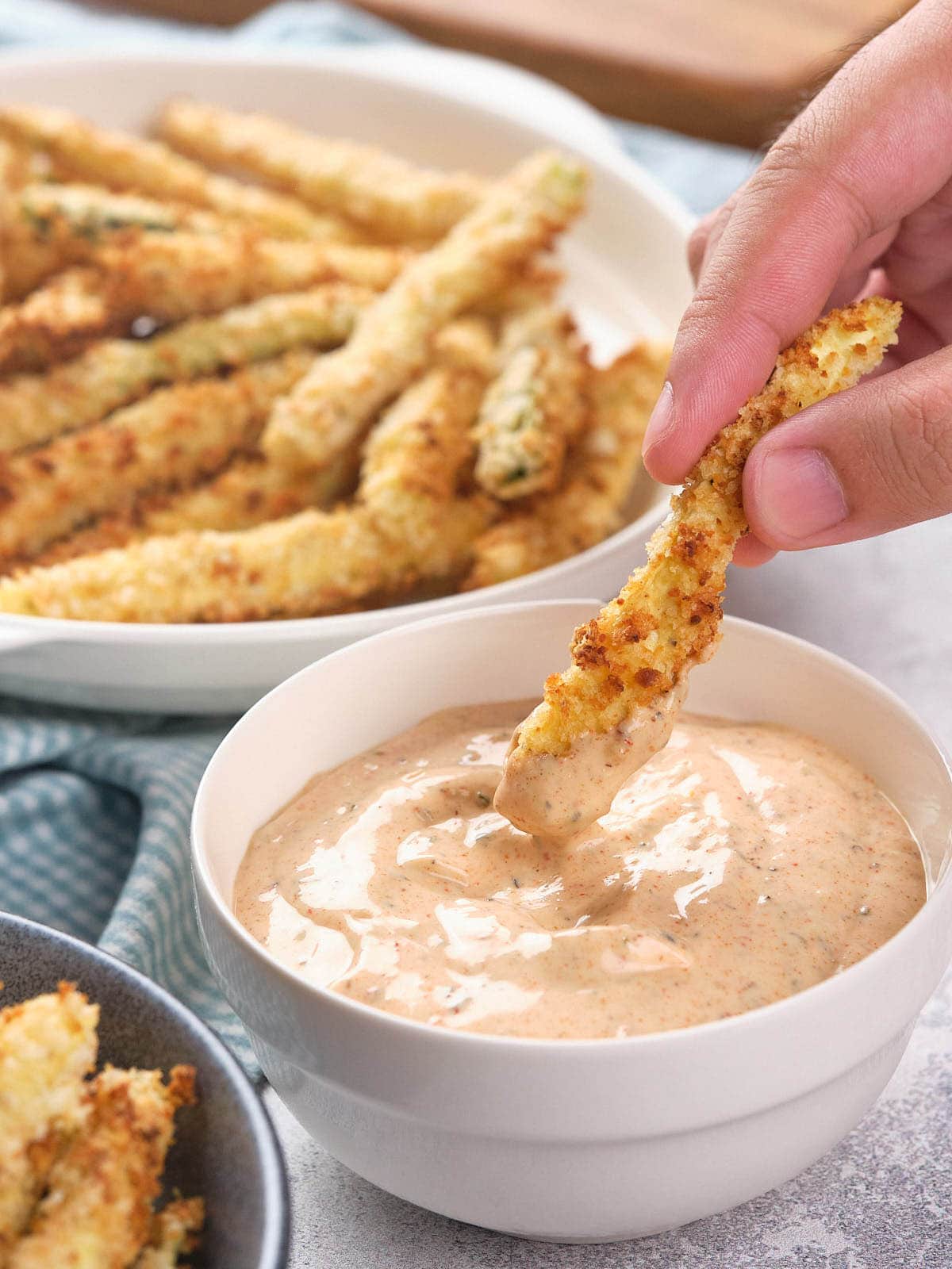 A hand dips a breaded zucchini fry into a bowl of creamy dipping sauce, with more zucchini fries on a plate in the background.