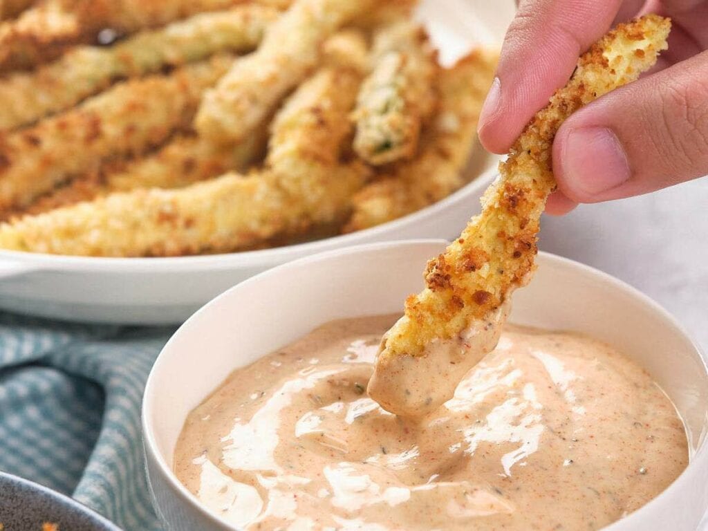 A hand dips a breaded zucchini fries into a bowl of creamy dipping sauce.