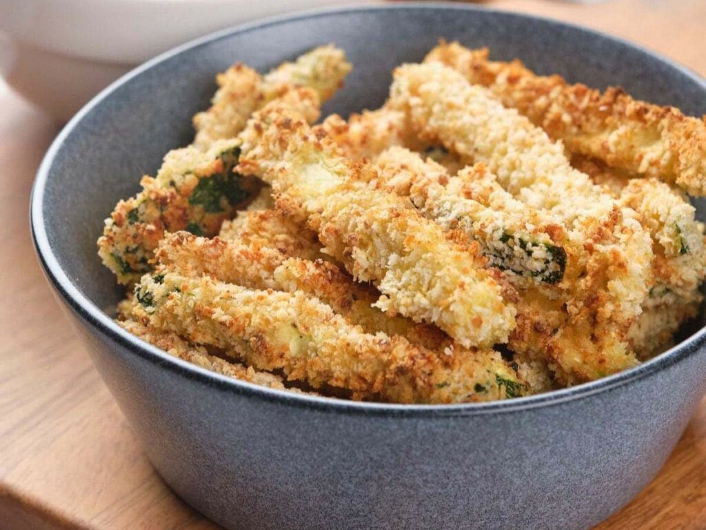 A gray bowl filled with breaded and baked zucchini fries on a wooden surface.